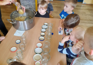 Dzieci siedzą wokół stołu, nauczycielka nalewa mus z garnka łyżką wazową